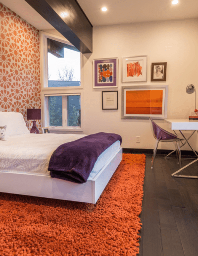 A modern bedroom with a white bed, vibrant orange carpet, decorative wallpaper, and a small white dog resting on the bed.