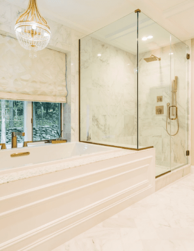 A spacious, well-lit bathroom featuring a large tub, glass shower enclosure, marble walls, and a chandelier, with a view of the outside through a window.