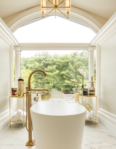 A modern bathroom features a freestanding white tub, gold fixtures, side tables with towels and toiletries, large arched window, and a view of greenery outside.