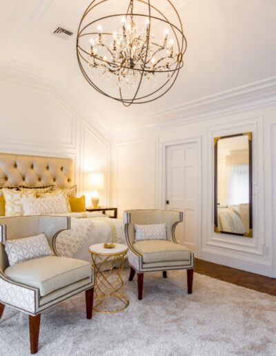 Elegant bedroom with a large bed, two armchairs, a round table, chandelier, white walls, and a plush rug on wooden flooring.